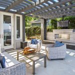 Modern outdoor patio area with wicker furniture, cushions, a beige dog bed, and a wall-mounted tv, under a wooden pergola adjacent to a sliding glass door.