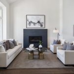 Modern living room with white sofas, a dark fireplace, abstract wall art, and large windows.