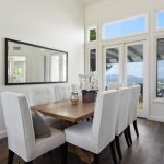 Modern dining room with a wooden table, white upholstered chairs, large windows showing a city view, and a wall mirror.
