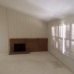 Empty living room with a brick fireplace, white walls, and large windows with closed blinds.