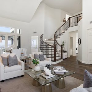 Bright, spacious living room with white sofas, a glass coffee table, dark hardwood floors, and a winding staircase leading to an upper level.