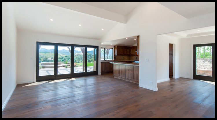 Empty modern living room with hardwood floors, white walls, and large windows overlooking a scenic view, with a wooden kitchen counter in the background.