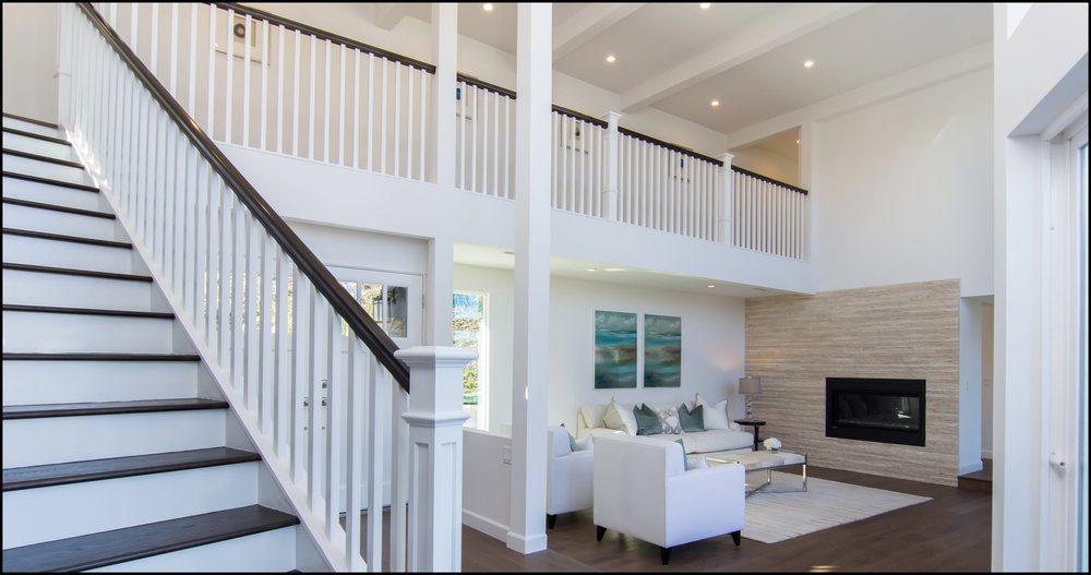 Modern living room with white sofas and a fireplace, flanked by a staircase leading to an upper balcony with railings, featuring spacious, bright interiors.
