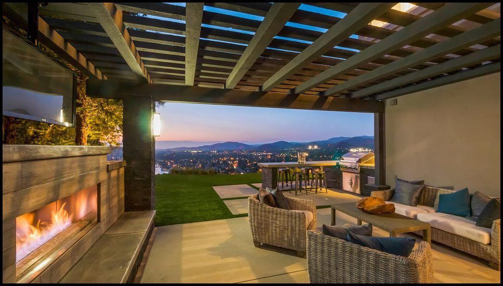 Modern outdoor patio with a fireplace, comfortable seating, and a panoramic view of a cityscape at dusk under a pergola.